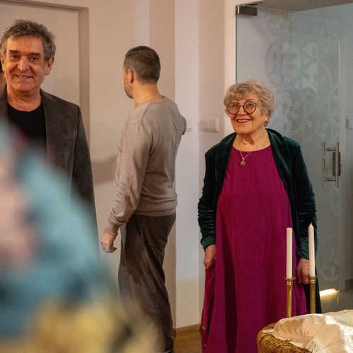 Uśmiechnięta autorka wystawy w fioletowej sukni oraz zielonym płaszczu stoi obok stołu z dwiema świecami.