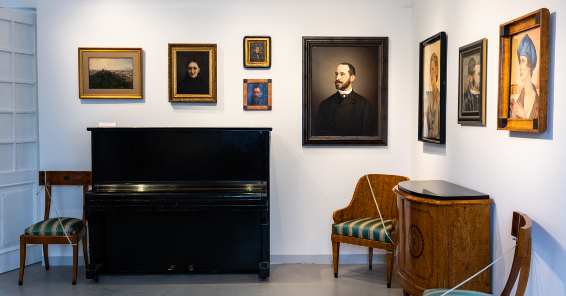 Widok na ściany z obrazami, z lewej strony pianino po prawej fotele i mała szafka