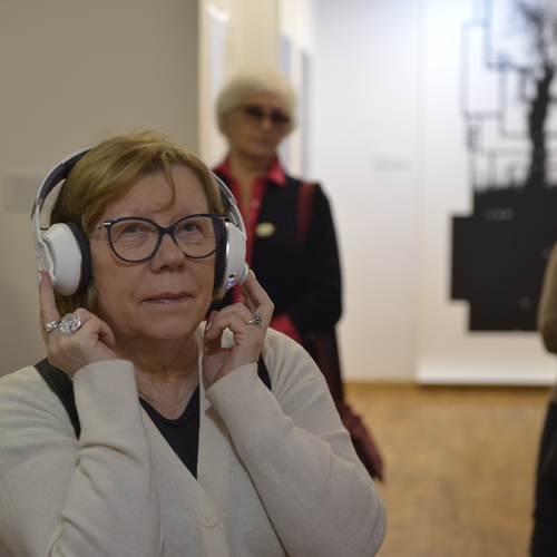 Starsza kobieta w okularach słucha nagrania przez słuchawki podczas zwiedzania wystawy w galerii.