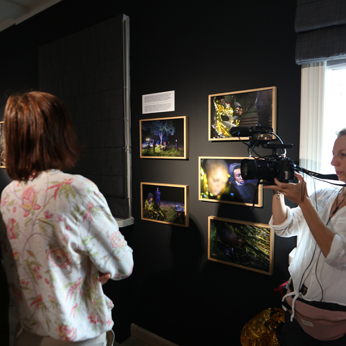 Dwie osoby oglądają fotografie na czarnej ścianie galerii, podczas gdy kobieta z kamerą nagrywa wystawę.