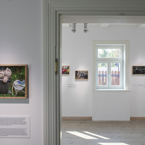 Widok dwóch jasnych pomieszczeń galerii z fotografiami dokumentującymi kryzys humanitarny, w tym zbliżenie na zdjęcie poranionych stóp spoczywających na walizce.