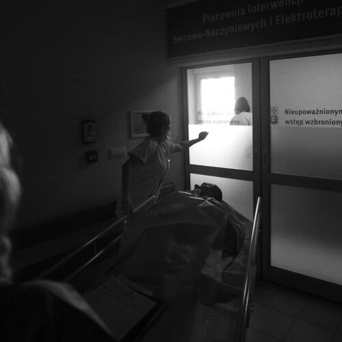 Czarno - biała fotografia reportażowa. Pielęgniarka w szpitalu wiezie pacjenta  na łóżku szpitalnym przez drzwi z napisem „Nieupoważnionym wstęp wzbroniony”.