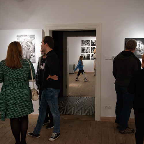 Zwiedzający oglądają czarno-białe fotografie na ścianach galerii, podczas gdy w oddali przez drzwi przechodzi dziecko w niebieskiej bluzie.