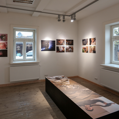 Wnętrze galerii z drewnianą podłogą, gdzie na ścianach wiszą kolorowe fotografie, a na środku pomieszczenia znajduje się podłużna ława z nadrukowanymi zdjęciami.